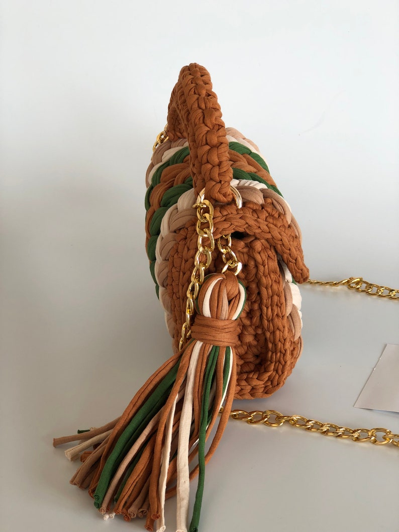 May include: A brown, green, and cream crocheted handbag with a gold chain strap and tassel.