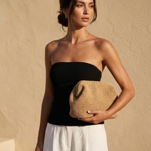 Puede incluir: Un bolso de mano tipo sobre hecho de paja tejida en color beige con una estructura r&iacute;gida y superficie texturizada. El bolso tiene forma rectangular con esquinas redondeadas y una abertura superior, sostenido horizontalmente sobre ropa clara.