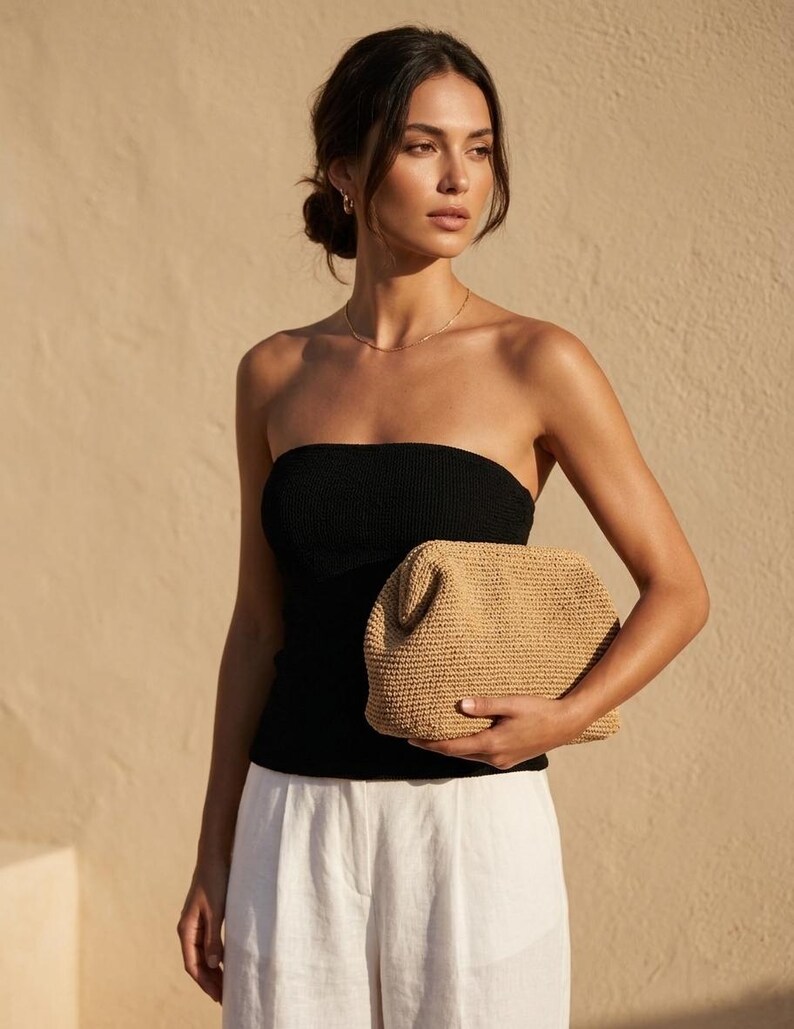 Puede incluir: Un bolso de mano tipo sobre hecho de paja tejida en color beige con una estructura r&iacute;gida y superficie texturizada. El bolso tiene forma rectangular con esquinas redondeadas y una abertura superior, sostenido horizontalmente sobre ropa clara.