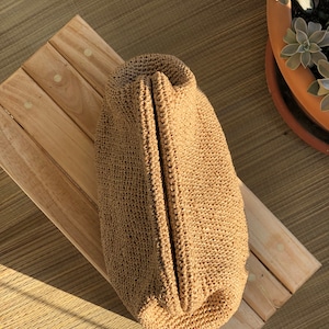 Puede incluir: Un bolso de mano tipo sobre tejido a ganchillo en color beige con una abertura de marco r&iacute;gido articulado. El bolso presenta un patr&oacute;n de puntadas denso y uniforme en un material similar a la fibra natural, con una forma alargada.