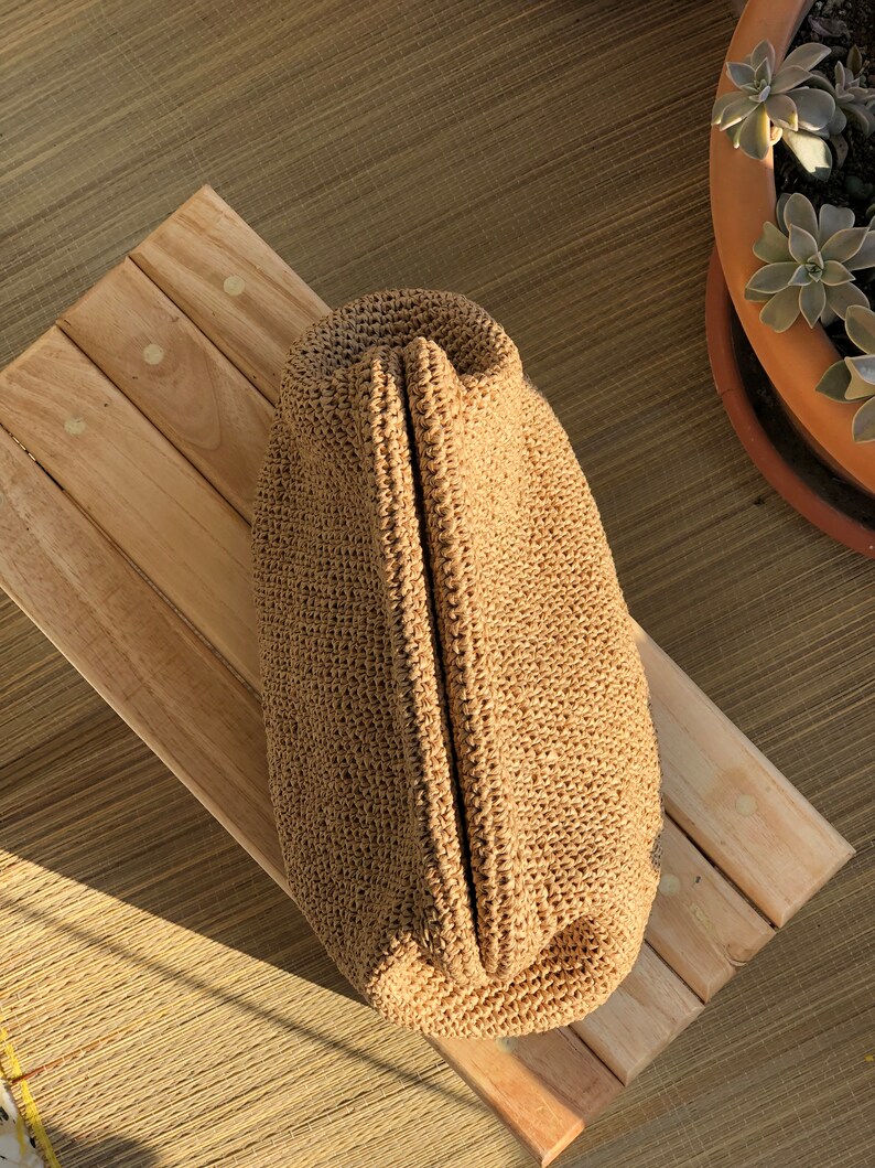 Puede incluir: Un bolso de mano tipo sobre tejido a ganchillo en color beige con una abertura de marco r&iacute;gido articulado. El bolso presenta un patr&oacute;n de puntadas denso y uniforme en un material similar a la fibra natural, con una forma alargada.