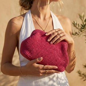 May include: A vibrant magenta clutch bag crafted from a textured, woven material. The bag features a rounded, soft-structured shape with a secure top closure. The surface displays a consistent, dense crochet-like pattern in a deep pink hue.