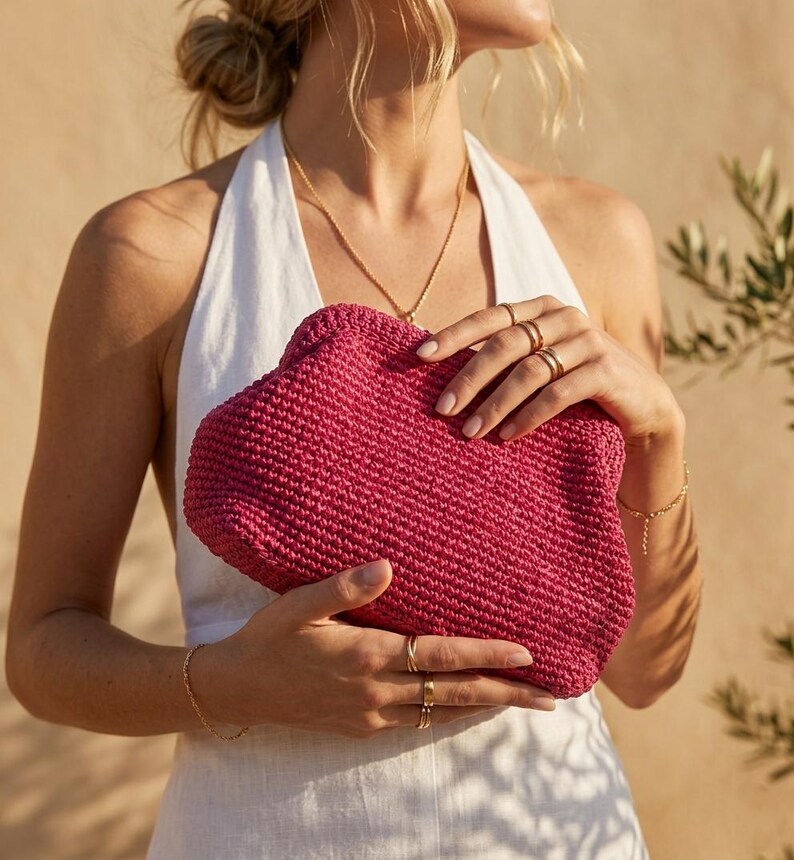 May include: A vibrant magenta clutch bag crafted from a textured, woven material. The bag features a rounded, soft-structured shape with a secure top closure. The surface displays a consistent, dense crochet-like pattern in a deep pink hue.
