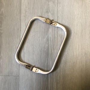 May include: Silver-toned metal bag handle with two gold-toned hinges. The handle is rectangular in shape and is designed to be attached to a bag.