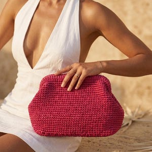 May include: A vibrant magenta clutch bag crafted with a dense, textured crochet knit pattern. The bag features a structured, rounded frame design with a secure top closure, showcasing a consistent, uniform stitch throughout the entire exterior.