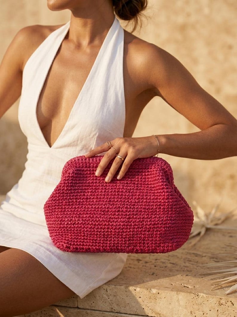 May include: A vibrant magenta clutch bag crafted with a dense, textured crochet knit pattern. The bag features a structured, rounded frame design with a secure top closure, showcasing a consistent, uniform stitch throughout the entire exterior.
