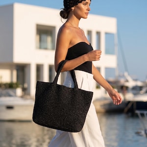 May include: A black woven tote bag with two sturdy handles, featuring a textured, grid-like pattern throughout the exterior. The bag has a rectangular shape and is constructed from a dark, fibrous material, providing a structured appearance.