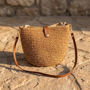 Op de afbeelding: Een beige gehaakte emmertas met een bruine lederachtige riem en een centrale verticale bruine lederachtige lip met een goudkleurige metalen drukknoopsluiting. De tas heeft een geweven textuur en is bevestigd met goudkleurige metalen clips.