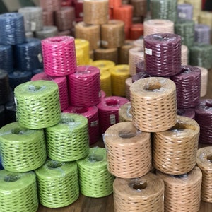 May include: Assortment of colorful paper twine spools in various colors, including green, pink, brown, and beige. The spools are stacked in a pyramid shape on a wooden table.