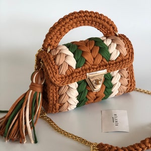 May include: A small, brown, green, and white crocheted handbag with a gold chain strap and a brown tassel. The bag has a gold clasp and the brand name "HILENIS" is printed on a white tag.