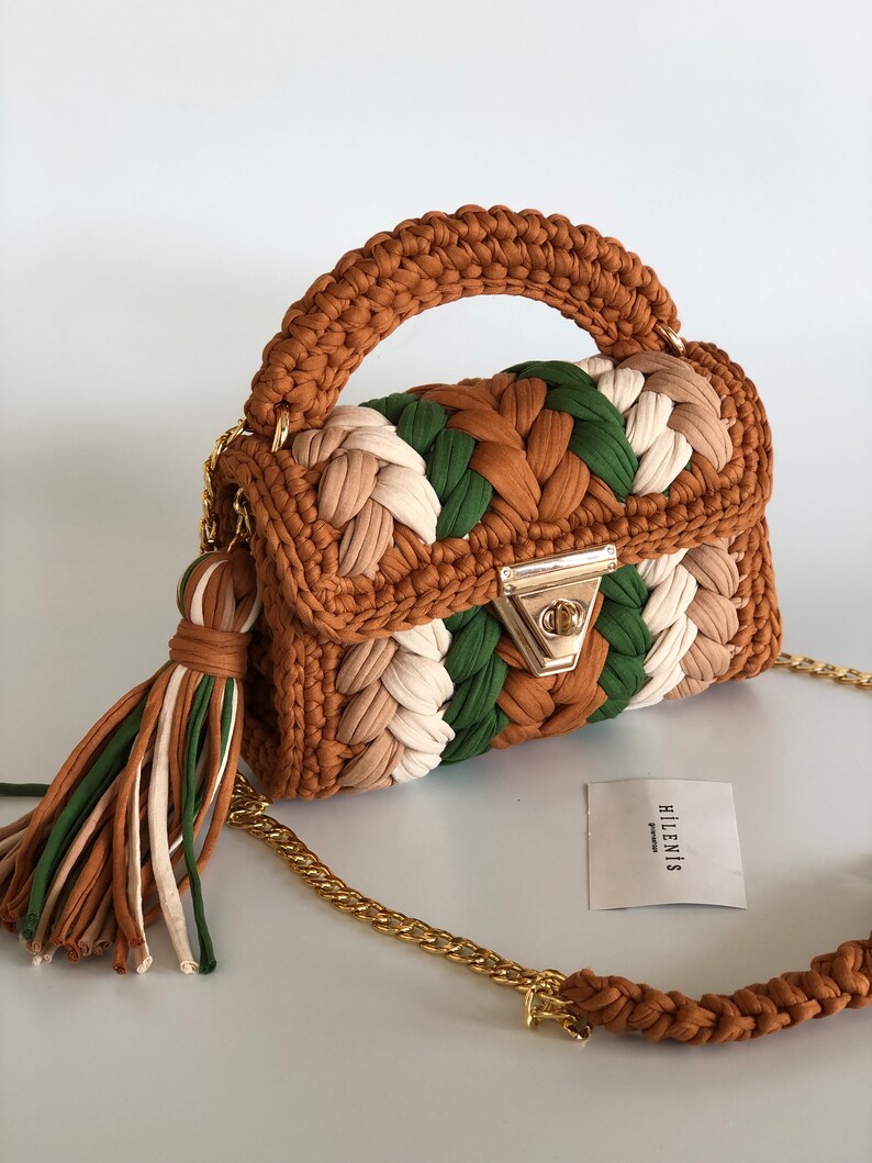 May include: A small, brown, green, and white crocheted handbag with a gold chain strap and a brown tassel. The bag has a gold clasp and the brand name "HILENIS" is printed on a white tag.
