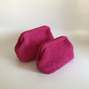 May include: Two fuchsia-colored crochet pouches with a structured frame opening. Each pouch features a dense, uniform stitch pattern and a rounded, compact shape. The bags are identical in color and texture, positioned side by side on a plain surface.