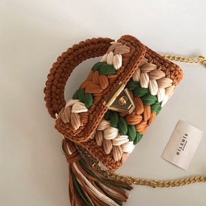 May include: A small brown, green, and cream crocheted handbag with a gold chain strap and tassel. The bag has a gold clasp and the brand name "HILLENIS" is printed on a white card.