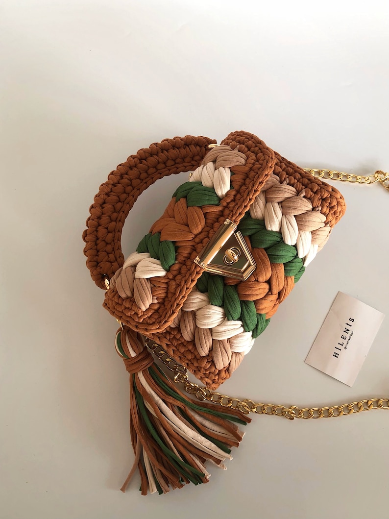May include: A small brown, green, and cream crocheted handbag with a gold chain strap and tassel. The bag has a gold clasp and the brand name "HILLENIS" is printed on a white card.