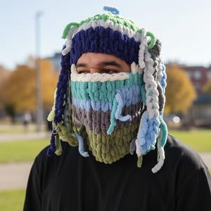 May include: A chunky knit balaclava crafted from thick, soft yarn in horizontal stripes of olive green, grey, light blue, and white. The headpiece features dangling braided yarn strands in shades of navy, light blue, and green, accented with small silver metal rings.
