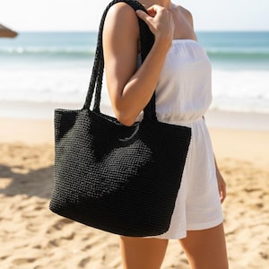 May include: A black tote bag with a textured, woven construction and two braided shoulder straps. The bag features a rectangular shape with a flat base and a consistent, dense stitch pattern throughout the exterior surface of the accessory.
