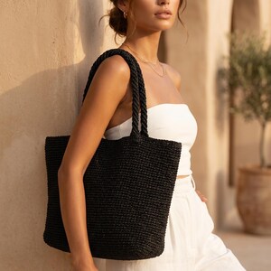 May include: A black woven tote bag with two sturdy braided handles. The bag features a rectangular shape and a textured, straw-like material construction. The design is simple and functional, providing a spacious interior compartment for carrying items.