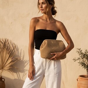 Puede incluir: Un bolso de mano tejido en color beige con una abertura de marco estructurado. El bolso presenta un exterior texturizado de ganchillo en un tono tostado neutro, una forma rectangular con esquinas redondeadas y un cierre superior articulado.
