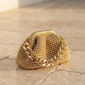 May include: A gold-colored clutch bag with a textured, woven exterior and a metallic frame closure. The bag features a thick, polished gold-tone chain handle that drapes across the front, providing a distinct contrast to the woven body.
