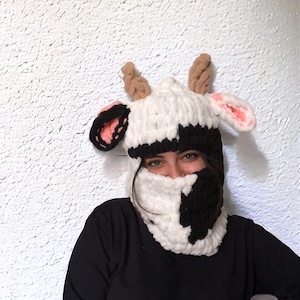 May include: A black and white cow-patterned knit hat and neck warmer with brown horns and pink ears.