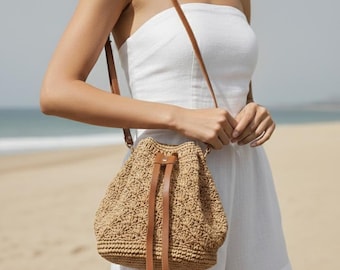 May include: A tan crochet bucket bag with a textured shell pattern, featuring a brown leather drawstring closure and a matching thin brown leather shoulder strap. The bag has a structured circular base and a soft, woven body construction.