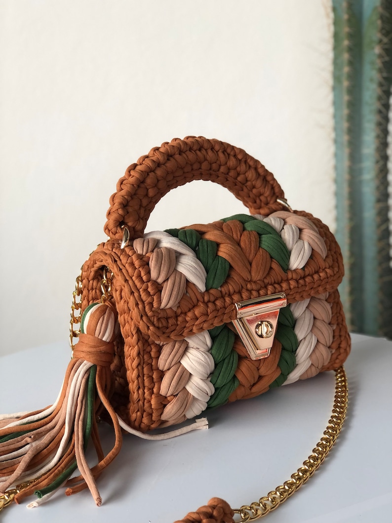 May include: A small brown, green, and white crocheted handbag with a gold chain strap and a tassel. The bag has a top handle and a gold clasp.