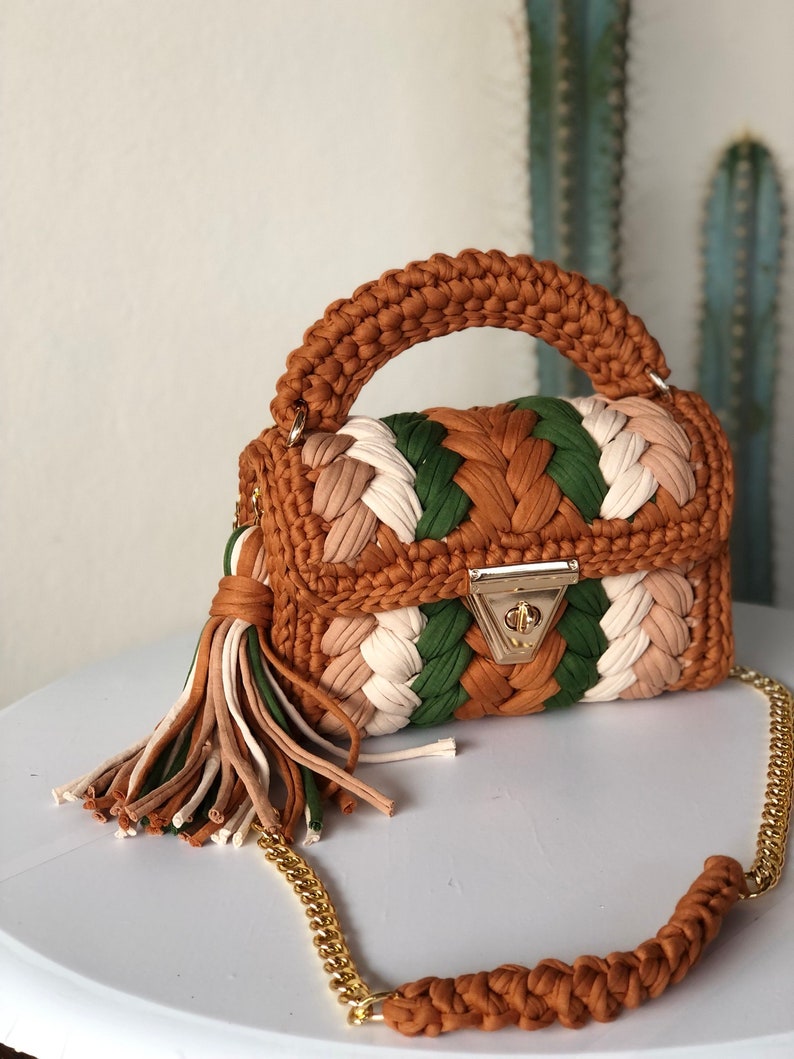 May include: A small, brown, green, and white crocheted handbag with a gold chain strap and a tassel. The bag has a woven design and a gold clasp.