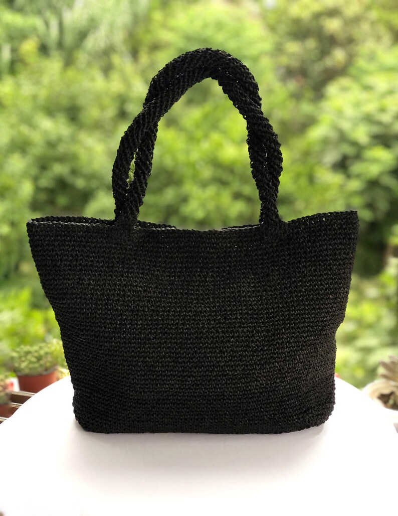 May include: A black tote bag constructed from a woven material with a textured surface. The bag features two sturdy, twisted handles attached to the top edge, providing a consistent dark aesthetic throughout the entire structure of the item.