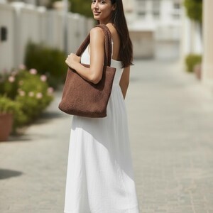 May include: A dark brown crocheted tote bag with two thick shoulder straps, featuring a dense, textured knit pattern. The bag is rectangular in shape and is carried over the shoulder, showcasing a solid, uniform dark brown color throughout.