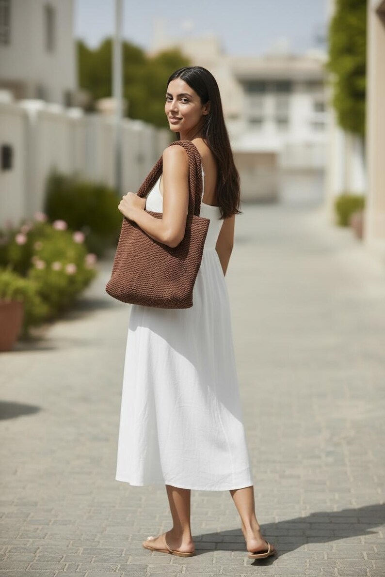 May include: A dark brown crocheted tote bag with two thick shoulder straps, featuring a dense, textured knit pattern. The bag is rectangular in shape and is carried over the shoulder, showcasing a solid, uniform dark brown color throughout.