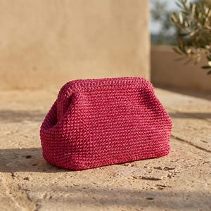 May include: A vibrant magenta clutch bag crafted with a dense, textured crochet stitch pattern. The accessory features a structured, rectangular frame with a kiss-lock closure mechanism, showcasing a consistent, uniform weave throughout the exterior.