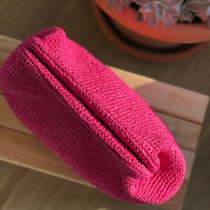 May include: A vibrant magenta pouch crafted from a dense, textured crochet knit. The rectangular accessory features a structured, folded top closure and a consistent, tight stitch pattern throughout its entire exterior surface.