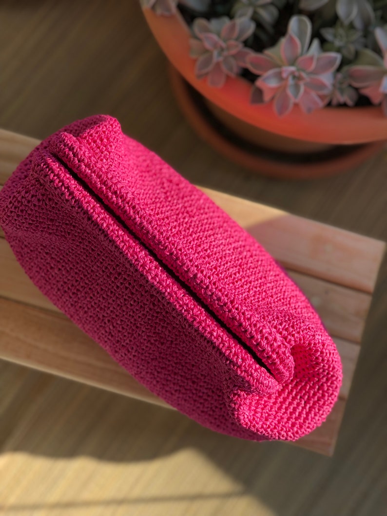 May include: A vibrant magenta pouch crafted from a dense, textured crochet knit. The rectangular accessory features a structured, folded top closure and a consistent, tight stitch pattern throughout its entire exterior surface.