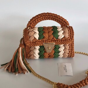 May include: A small, brown, green, and white crocheted handbag with a gold chain strap and a tassel. The bag has a gold clasp and the brand name "HILLENIS" is printed on a white card.