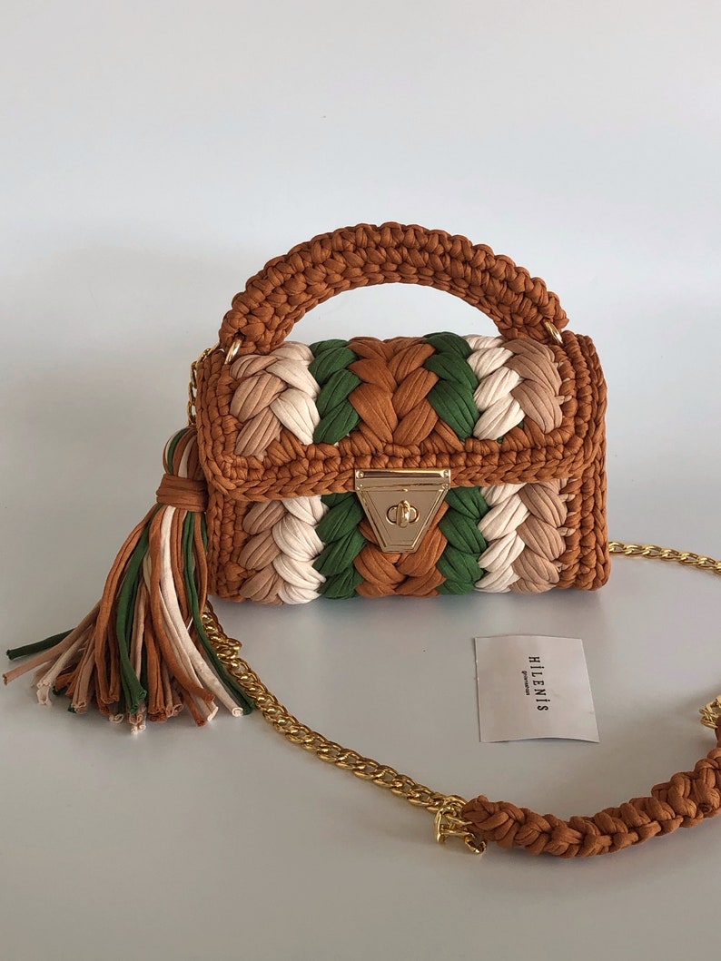 May include: A small, brown, green, and white crocheted handbag with a gold chain strap and a tassel. The bag has a gold clasp and the brand name "HILLENIS" is printed on a white card.