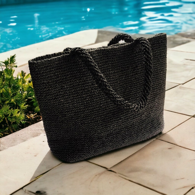 May include: A dark charcoal gray tote bag crafted from a woven, textured material. The bag features a rectangular silhouette with a flat base and two sturdy, braided handles attached to the top edge, providing a structured and functional design.