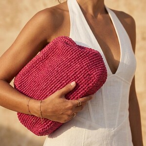 May include: A vibrant magenta clutch bag crafted from a thick, woven material with a rectangular, boxy shape. The bag features a textured, knotted surface pattern and a simple, clean design, held securely against the side of the body.