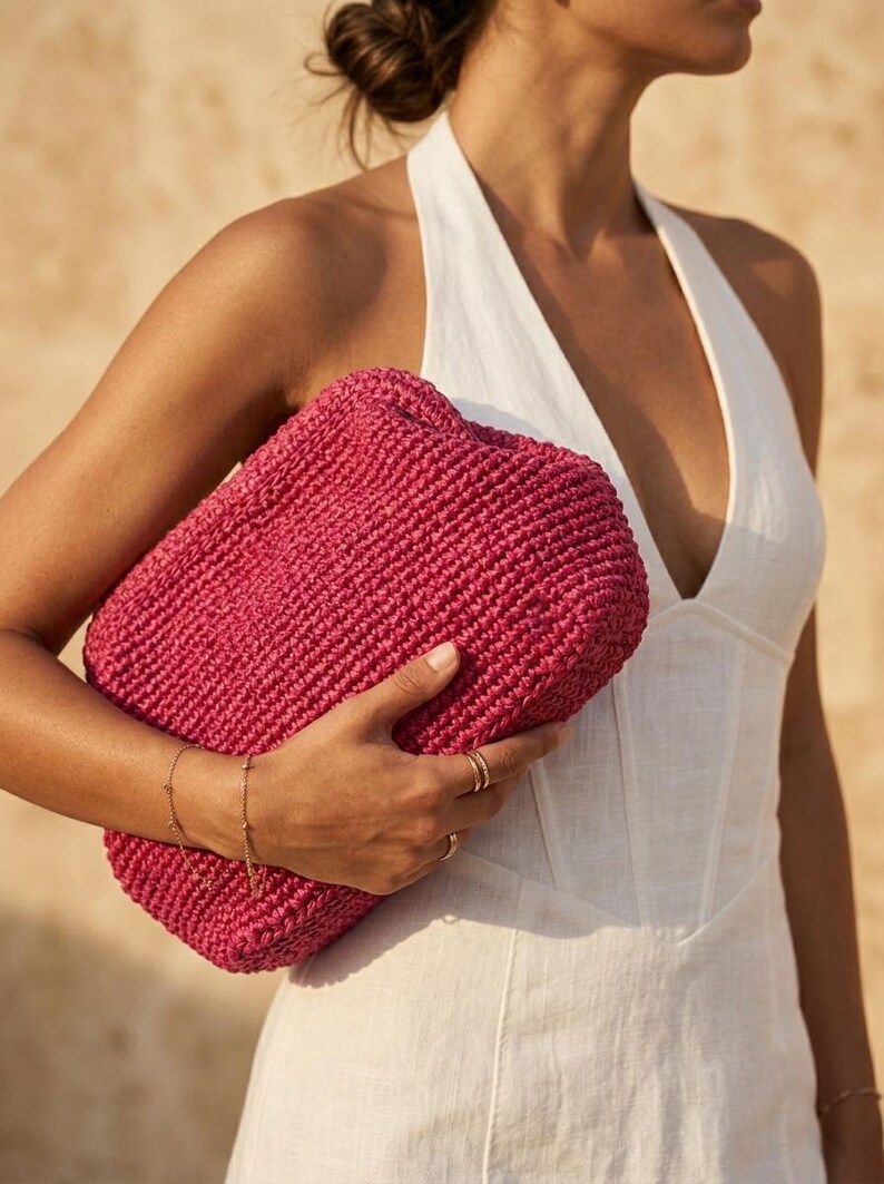 May include: A vibrant magenta clutch bag crafted from a thick, woven material with a rectangular, boxy shape. The bag features a textured, knotted surface pattern and a simple, clean design, held securely against the side of the body.