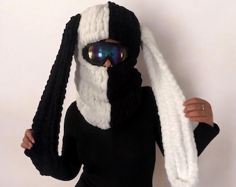 May include: A chunky knit balaclava featuring a split black and white color scheme with long, dangling rabbit-style ears. The headgear includes a large cutout for the eyes, which are covered by iridescent, dark-tinted ski goggles.