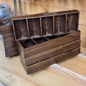 May include: A set of handcrafted wooden storage boxes with a rich, dark brown finish. The boxes feature multiple compartments, with a stacked design. The wood grain is visible, adding to the natural aesthetic. The boxes are designed for organizing small items.