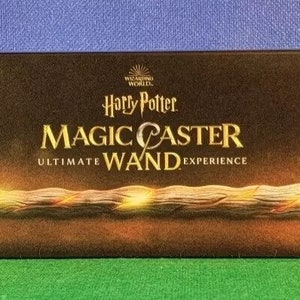 May include: A rectangular box with the words "Harry Potter Magic Caster Ultimate Wand Experience" in gold lettering. The box features a glowing wand with a textured handle and a bright orb at the end. The word "LOYAL" is in the lower left corner.