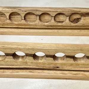 May include: Two wooden racks, each with a series of circular holes. The racks are made of light-colored wood with dark brown grain patterns. The top rack has six holes, while the bottom rack has seven. Both are rectangular with a base.