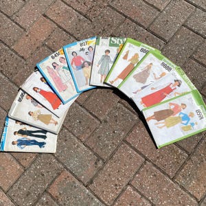 May include: A collection of ten vintage sewing pattern envelopes arranged in a fan shape. The envelopes feature various illustrations of clothing designs, including dresses, skirts, and trousers, with printed brand names like Simplicity and Style.