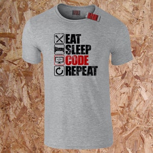 May include: A grey t-shirt featuring the text "EAT SLEEP CODE REPEAT" in black and red. The design incorporates icons of a knife and fork, a bed, a computer monitor, and a circular arrow. A barcode tag is attached to the shirt.