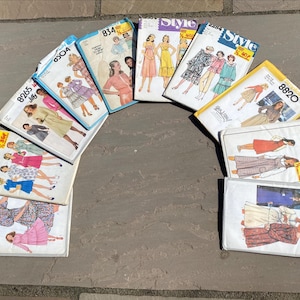 Puede incluir: Una colección de diez sobres de patrones de costura vintage dispuestos en semicírculo. Los sobres presentan varias ilustraciones de vestidos y conjuntos, con números de modelo impresos que incluyen 8265, 834, 8820, 2453, 8242 y 8383, junto a marcas como Simplicity y Style.
