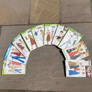 May include: A collection of ten vintage sewing pattern envelopes from Simplicity and Style brands, arranged in a semi-circle. Each white paper envelope features colourful illustrations of various clothing designs, including dresses, skirts, and trousers, with printed model numbers.