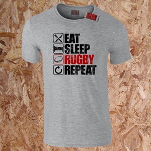 May include: A grey t-shirt featuring the text "EAT SLEEP RUGBY REPEAT" in black and red, with related icons. The design includes a crossed knife and fork, a bed, a rugby ball, and a circular arrow. A barcode tag is attached.