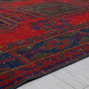 May include: A rectangular area rug featuring a vibrant red base color with intricate geometric patterns in deep blue and forest green. The design includes repeating central motifs and a wide, detailed border that frames the entire piece.