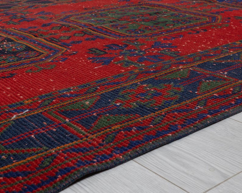May include: A rectangular area rug featuring a vibrant red base color with intricate geometric patterns in deep blue and forest green. The design includes repeating central motifs and a wide, detailed border that frames the entire piece.
