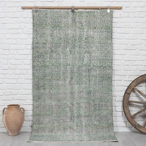 May include: A rectangular area rug featuring a faded, intricate floral pattern in shades of sage green, muted blue, and soft grey. The rug is suspended from a wooden rod by metal clips, displaying a weathered texture and a symmetrical design.